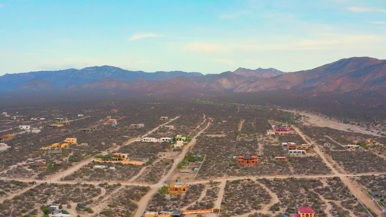 Aerial view over El Sargento La Ventana, Mexico, showing colorful houses among dry brush, intersecting dirt roads, a pale wash, distant ridges, and no visible people.