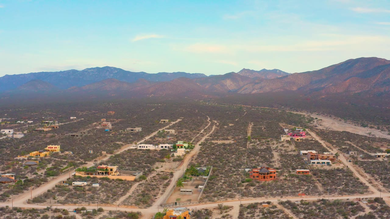 The aerial frame shows colorful buildings and dirt access roads in El Sargento La Ventana, with brown mountains in the background and no visible people.