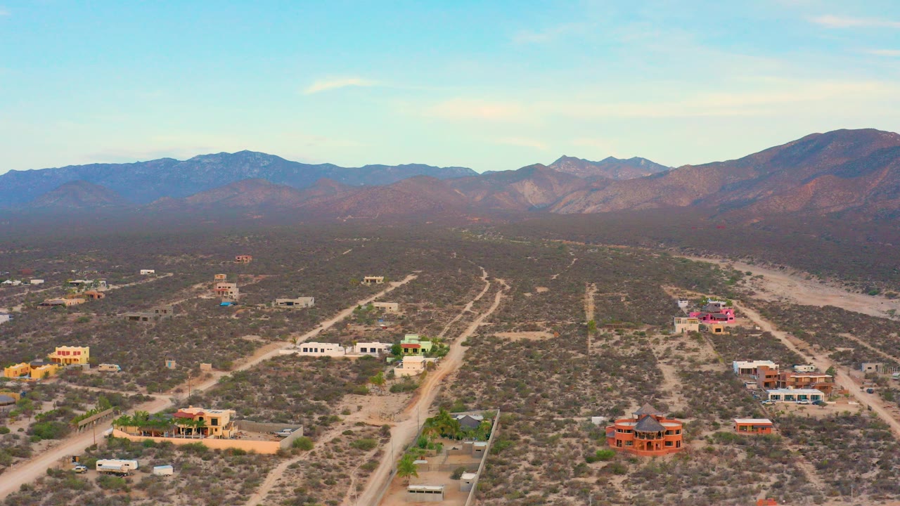 Opening aerial frame over El Sargento La Ventana, Mexico, with scattered desert buildings, dirt roads, a hazy mountain horizon, and no visible people.