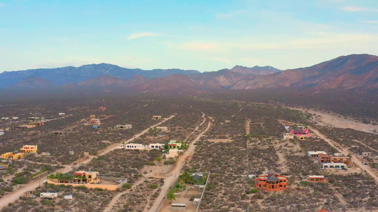 Aerial frame over El Sargento La Ventana showing scrubland lots, pale road lines, colorful low buildings, a dry wash to the right, and no visible people.