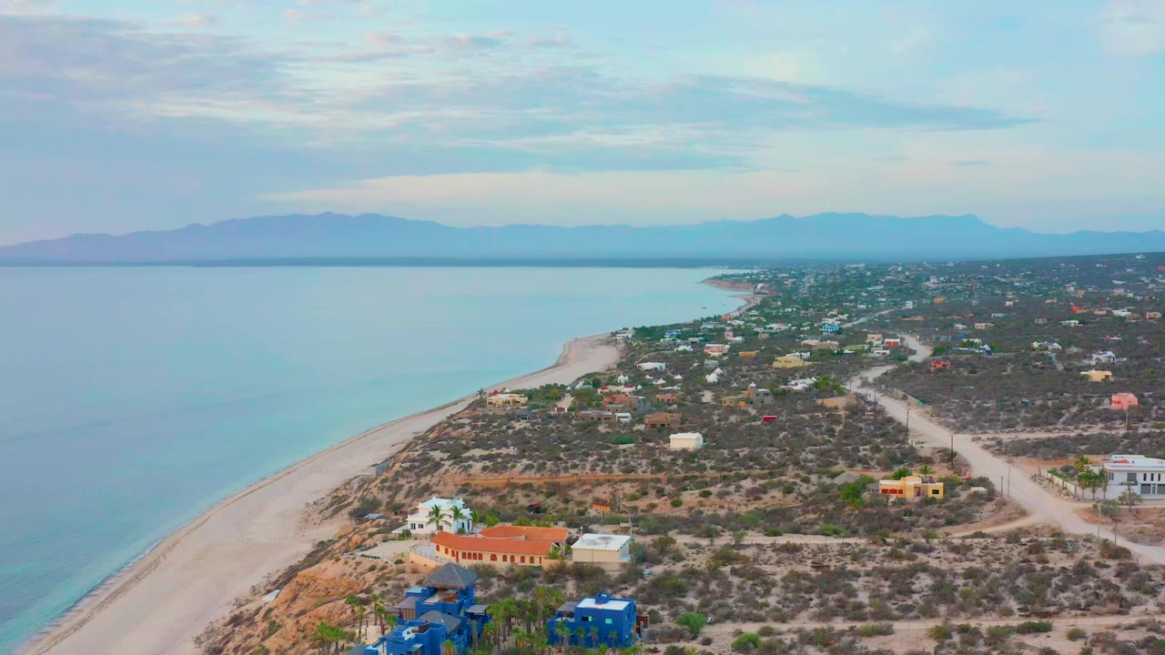 Early aerial frame over El Sargento La Ventana, Mexico, showing a pale beach curving beside turquoise water, scattered coastal buildings, dry foliage, distant mountains, and no visible people.