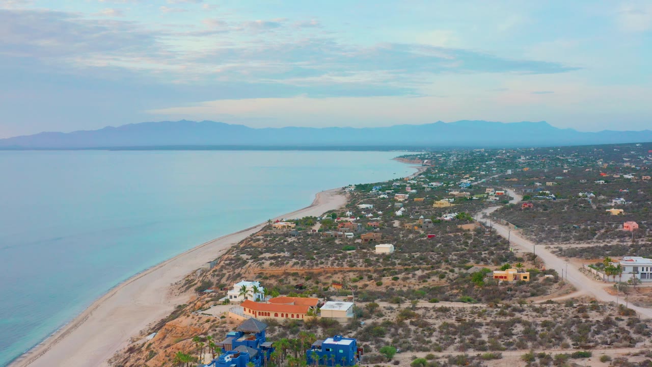 Wide aerial coastline with turquoise sea, pale beach, dry vegetation, buildings, and mountains on the horizon.
