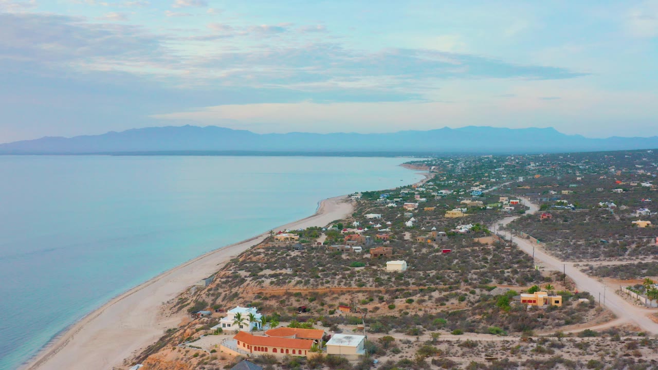 Opening aerial frame over El Sargento La Ventana, Mexico, with pale blue water at left, a sandy beach curve, scattered homes, dry scrubland, distant mountains, and no visible people.