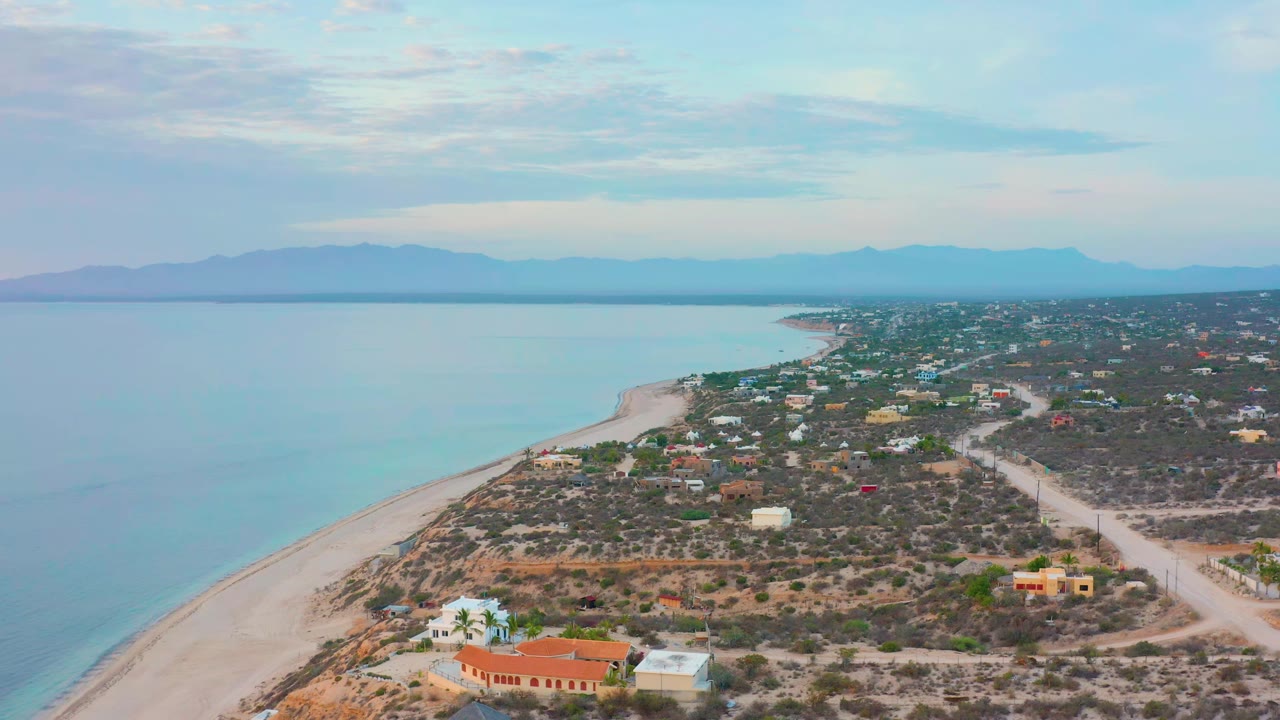 Wide aerial coastline with beach, low houses, scrubland, and mountains across the water.