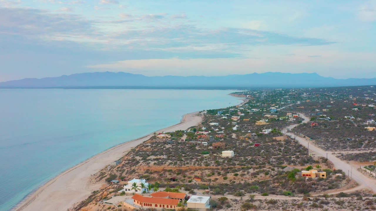 Aerial frame over El Sargento La Ventana showing clear water along the left, a narrow beach, low coastal homes, dirt tracks through scrub, hazy mountains, and no visible people.