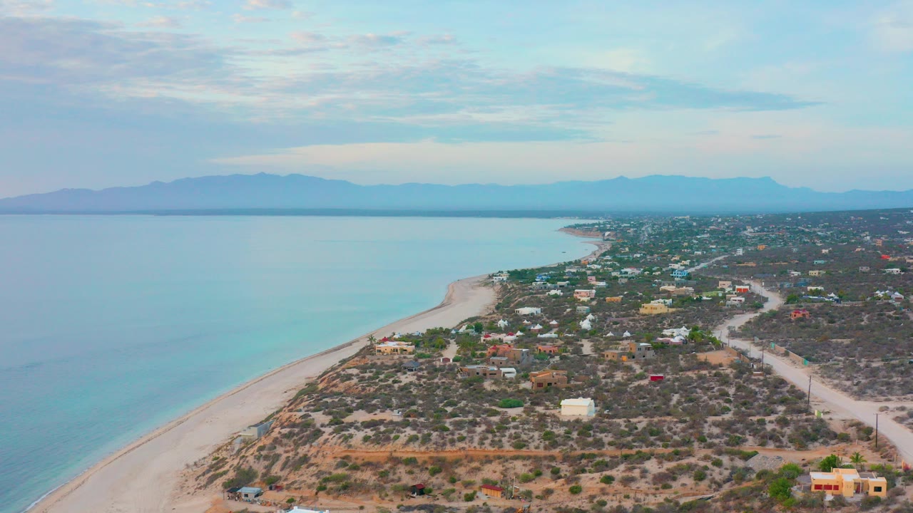 Opening aerial frame over El Sargento La Ventana, Mexico, with pale beach and blue-green water on the left, scattered homes and scrubland on the right, distant mountains, and no visible people.