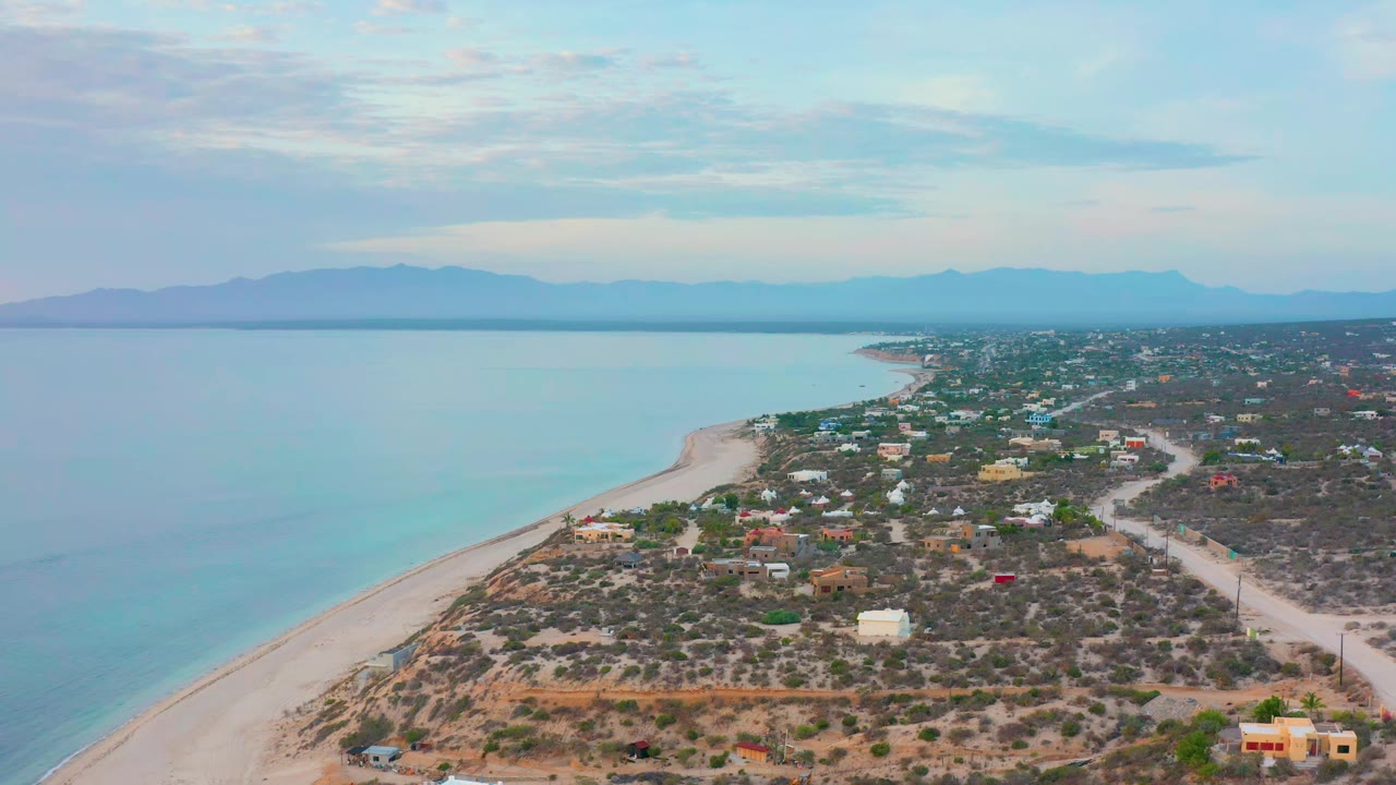 Second aerial frame over El Sargento La Ventana, Mexico, showing the beach curve slightly farther along the coast, with homes and dirt roads inland and no visible people.