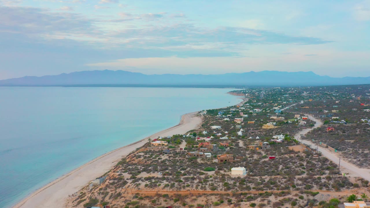 Wide aerial of coastline and residential scrubland with blue water to the left and mountains beyond.