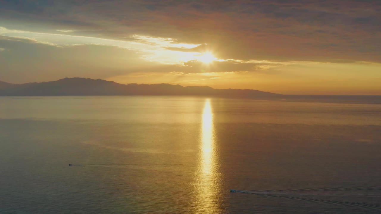 At 0:01, the sun reflection forms a vertical gold column on the ocean in El Sargento La Ventana, Mexico, with cloud cover above, a silhouetted coast behind it, small boats low in the frame, and no visible people.