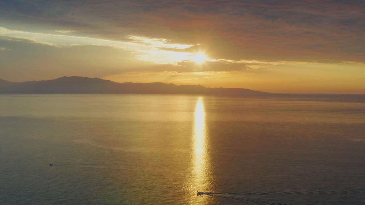 Low sun over calm water in El Sargento La Ventana, Mexico, with a bright gold reflection, distant mountain silhouettes, layered clouds, and no visible people.