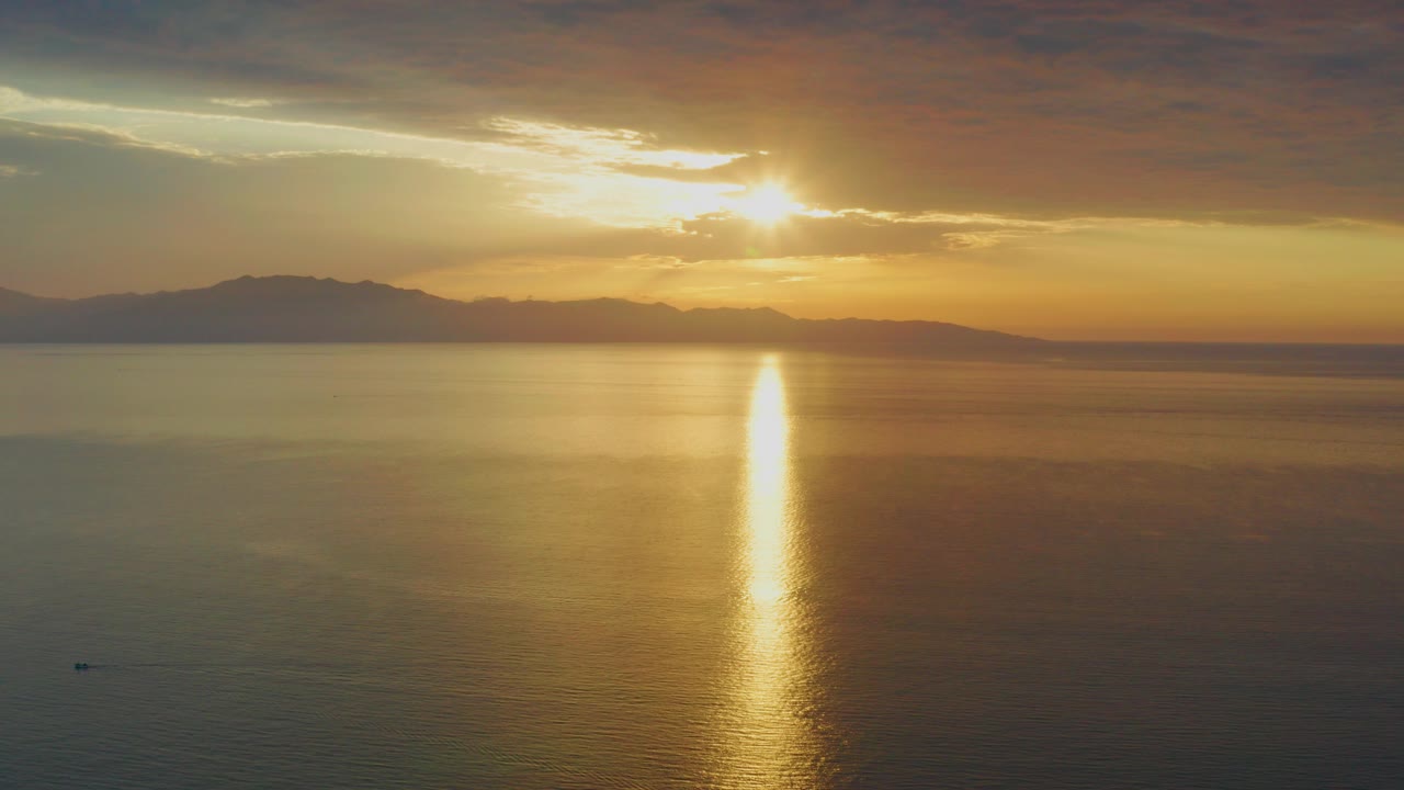 Opening frame of a sunset over the ocean at El Sargento La Ventana, Mexico, with the sun near a cloud band, a bright vertical reflection on the water, distant land on the horizon, and no visible people.