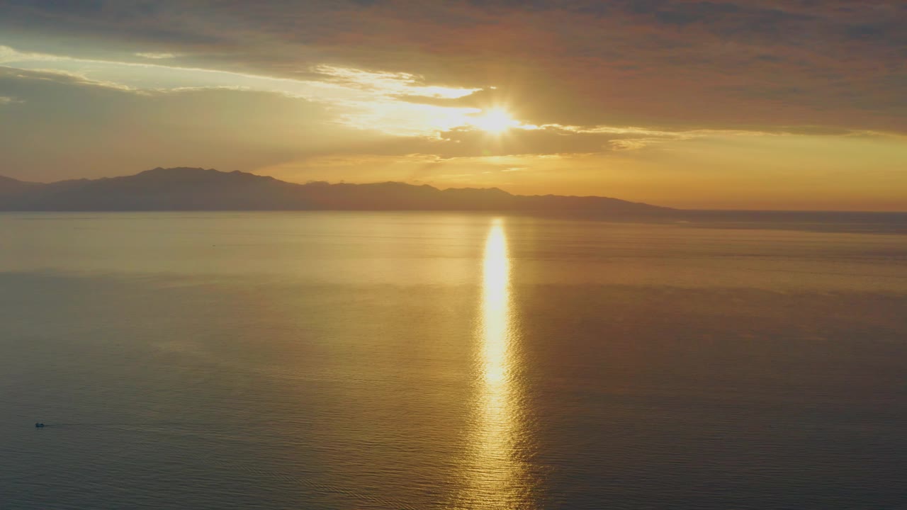 The sun sits just above the horizon cloud layer over El Sargento La Ventana, Mexico, with distant land stretching left of frame, gold light on the water, and no visible people.
