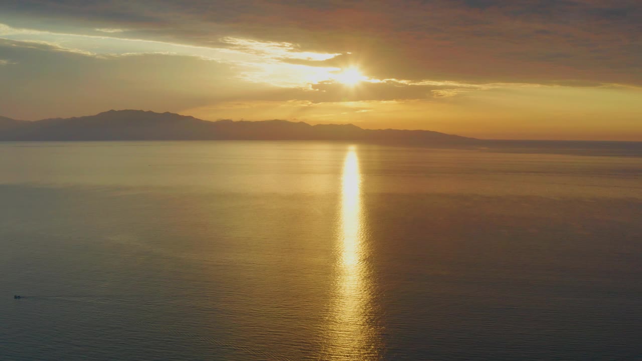 Mid-clip frame of the El Sargento La Ventana, Mexico sunset, with a dark cloud ceiling, orange sky near the horizon, far coastline, and a bright gold reflection on the ocean with no visible people.