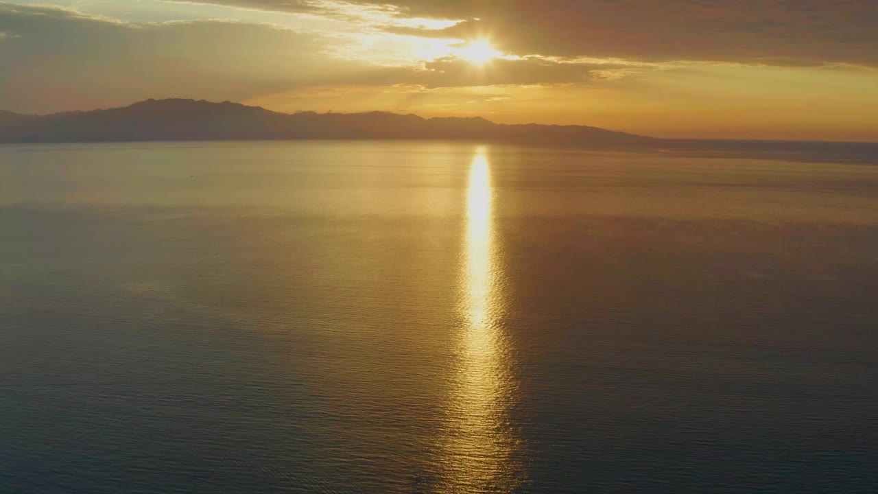 At 0:01, the sun remains partly framed by clouds above the mountain line, with the gold reflection falling near the center of the calm sea and no visible people.