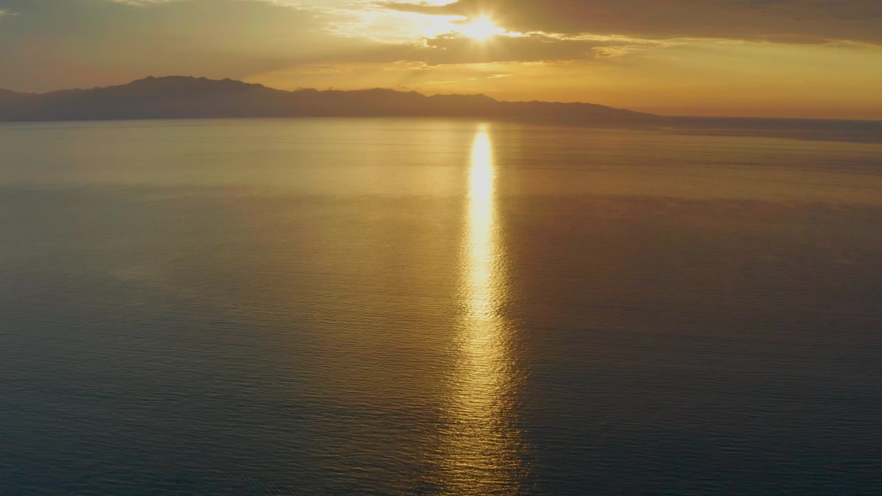 At 0:03, the sun sits near the top edge of the frame, the mountain ridge runs across the horizon, and a narrow gold reflection drops into the darker sea with no visible people.