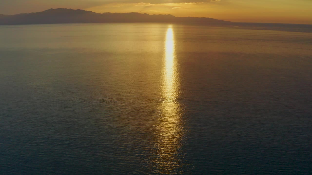 Opening frame of El Sargento La Ventana, Mexico at sunset, with a narrow golden reflection crossing dark calm water below a distant land silhouette and no visible people.