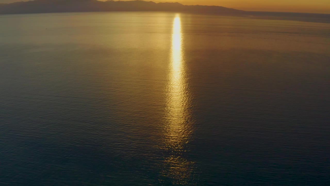 At 0:02, the aerial view shows a narrow bright column of sunset reflection reaching into darker foreground water, with the El Sargento La Ventana mountain horizon and no visible people.