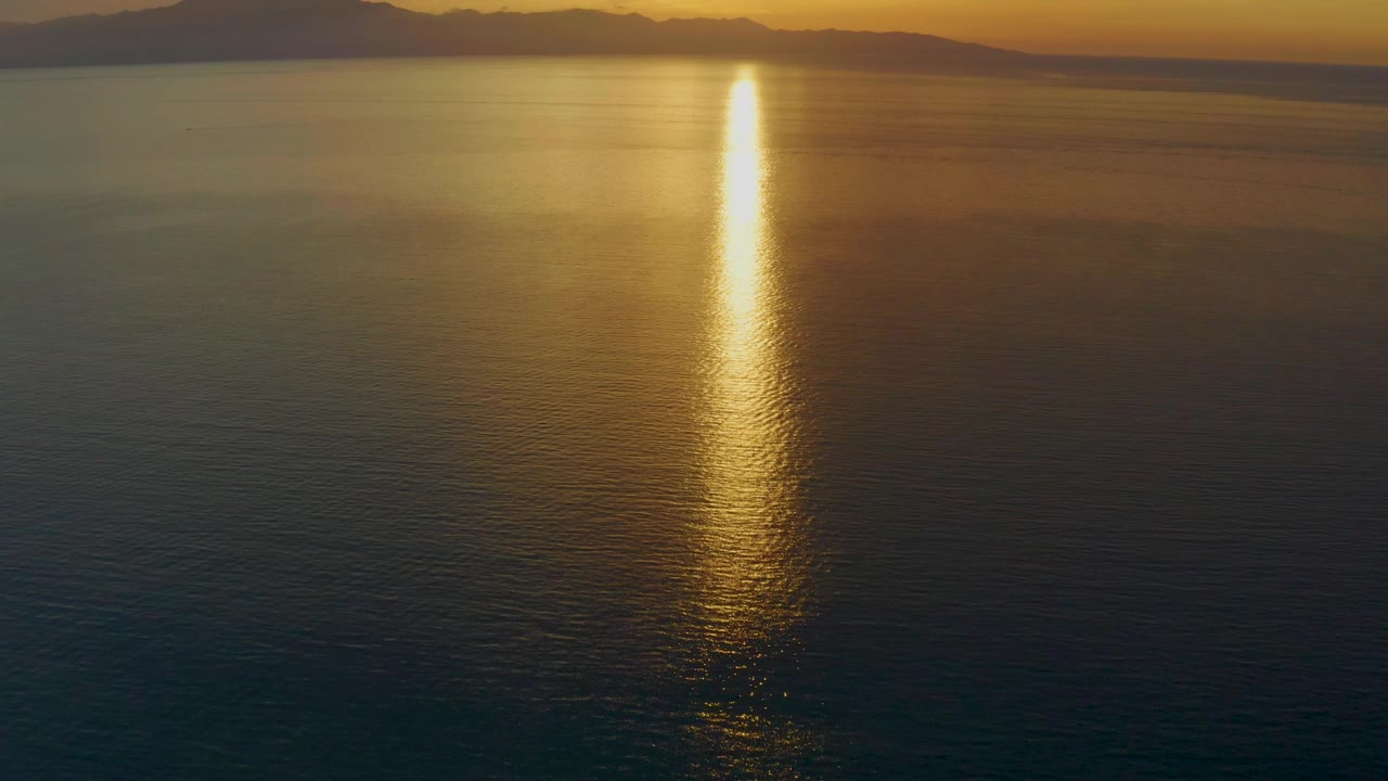 At 0:03, the composition stays high over El Sargento La Ventana water, with a bright golden reflection near the center, soft ripples, a dark mountain line, and no visible people.