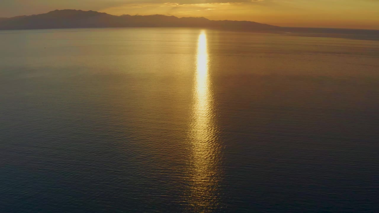 At 0:01, the golden reflection appears bright and narrow on the ocean, with darker water in the foreground and a low mountain ridge across the horizon in El Sargento La Ventana, Mexico, with no visible people.