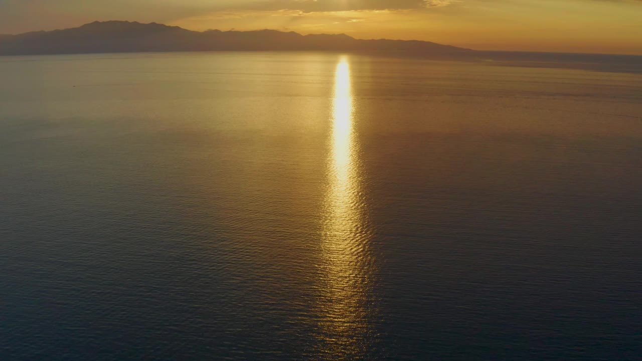 At 0:02, the sun reflection sits slightly right of center on calm water, with amber light spreading across the sea and silhouetted mountains behind it in El Sargento La Ventana, Mexico, with no visible people.