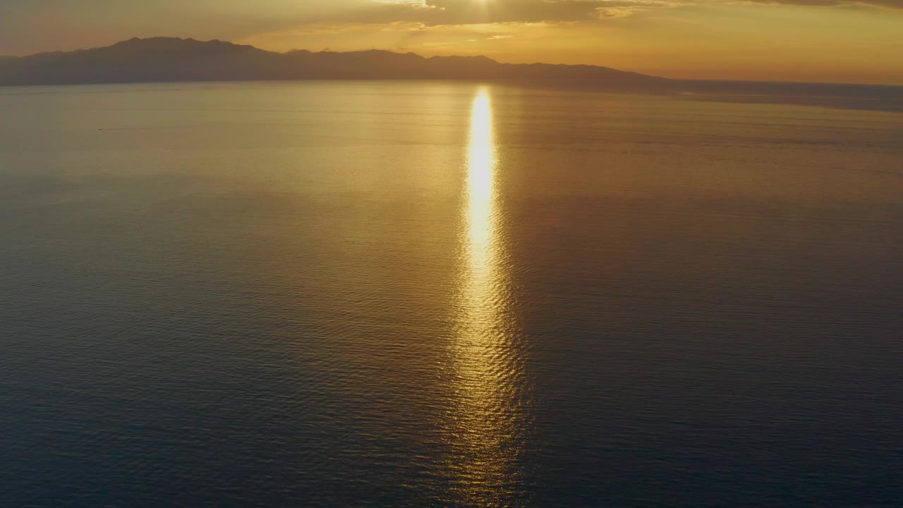 At 0:03, a wide aerial view over calm ocean shows a strong gold reflection reaching toward the camera, with distant mountains and a warm sky in El Sargento La Ventana, Mexico, with no visible people.