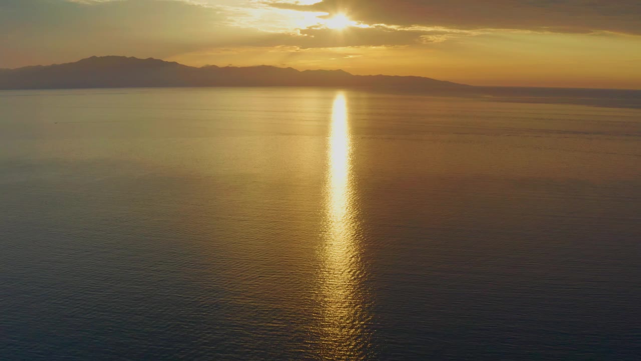 At 0:02 in El Sargento La Ventana, Mexico, amber sky spreads over a calm sea, with mountain silhouettes across the horizon and no visible people.