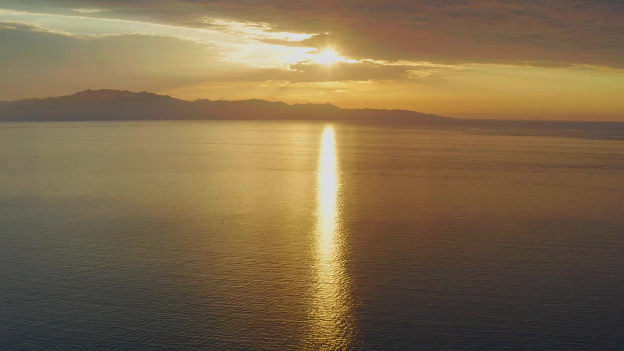A wide sunset frame in El Sargento La Ventana, Mexico, shows calm water, silhouetted mountains, warm clouds, and a bright reflection column with no visible people.