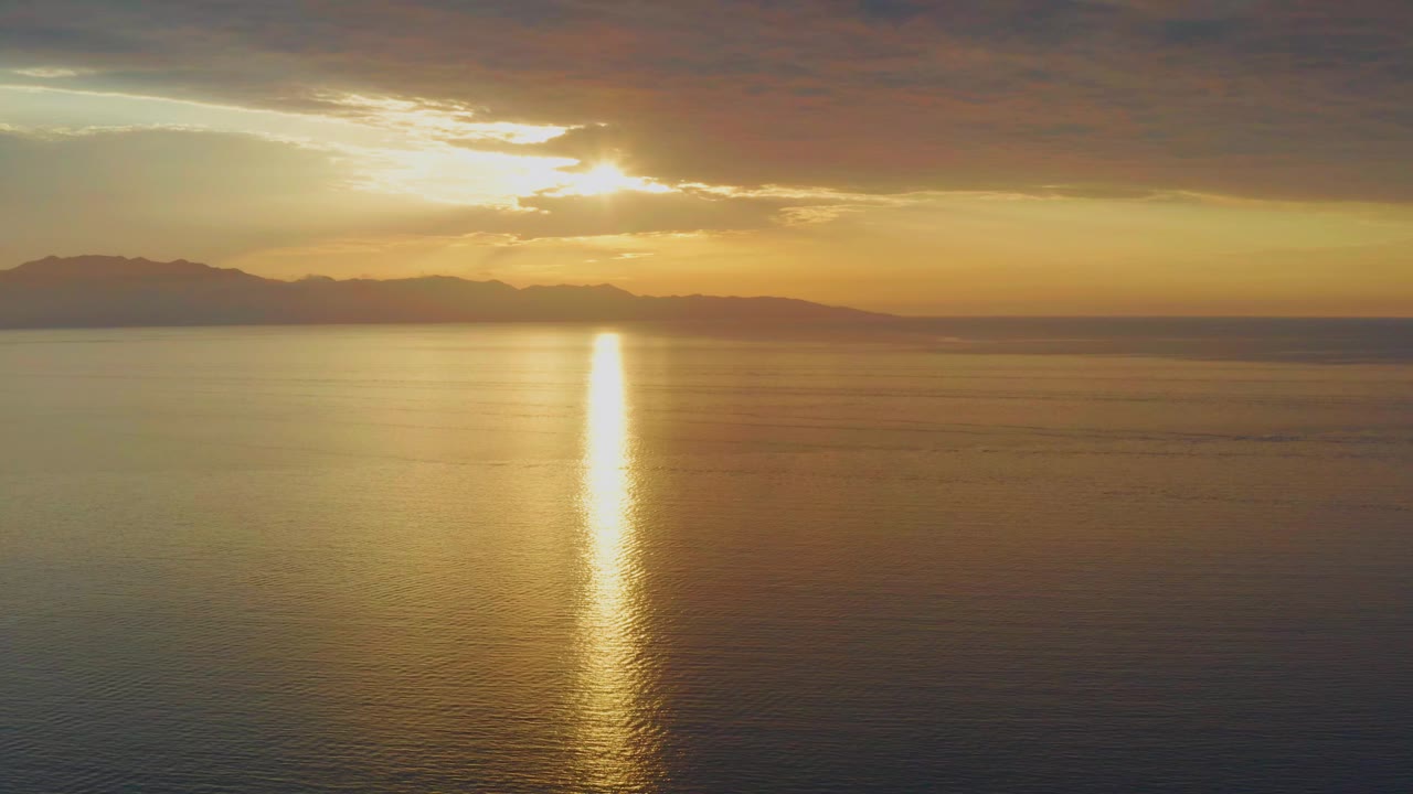 Opening frame of a golden sunset over the ocean in El Sargento La Ventana, Mexico, with the sun partly hidden by clouds, a bright reflection on the water, distant mountain silhouettes, and no visible people.