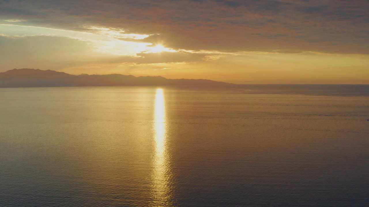 Opening frame of the El Sargento La Ventana sunset, with the sun breaking through clouds above distant mountains and a vertical gold reflection on calm water, no visible people.