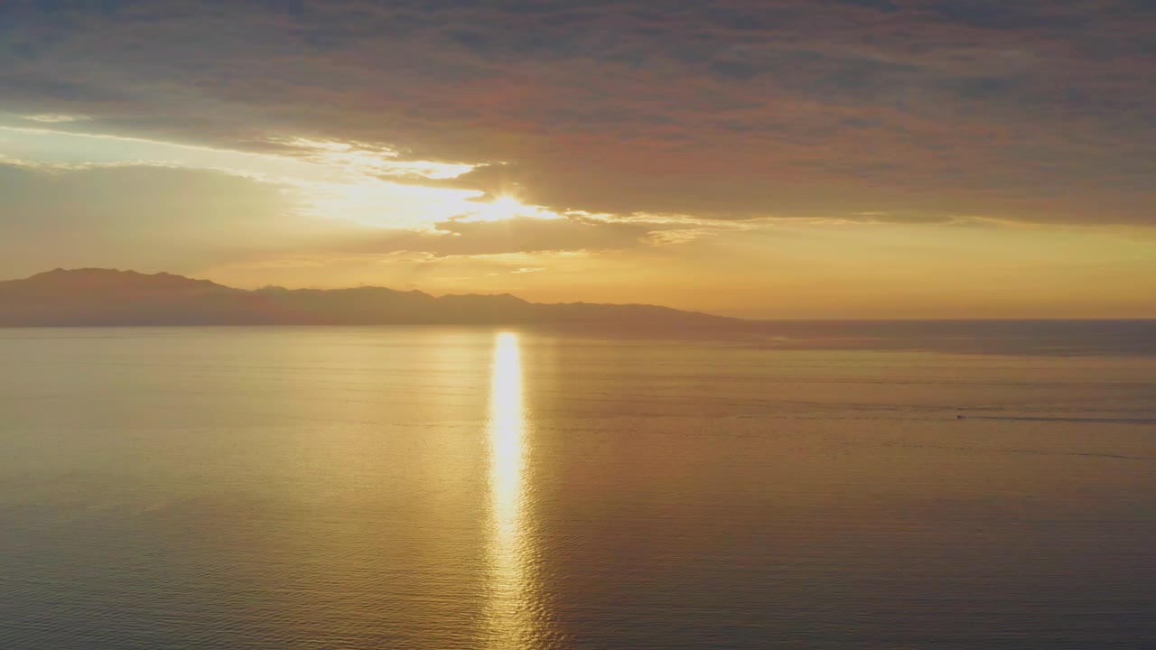 At 0:01, the golden reflection remains centered on the sea while a dark cloud band stretches across the top of the frame above the distant horizon in El Sargento La Ventana, Mexico, with no visible people.