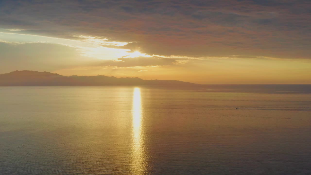 At 0:01, the sun sits behind a bright cloud opening over El Sargento La Ventana water, casting a narrow gold path across the sea toward a low mountain horizon, with no visible people.