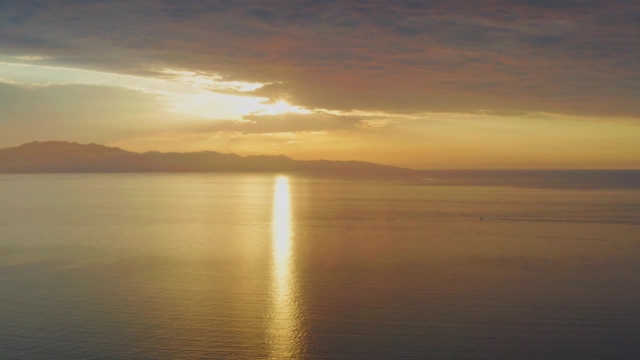 At 0:02, the wide 4K frame holds a golden sea surface, a bright vertical reflection below the hidden sun, layered clouds above, distant mountains to the left, and no visible people.