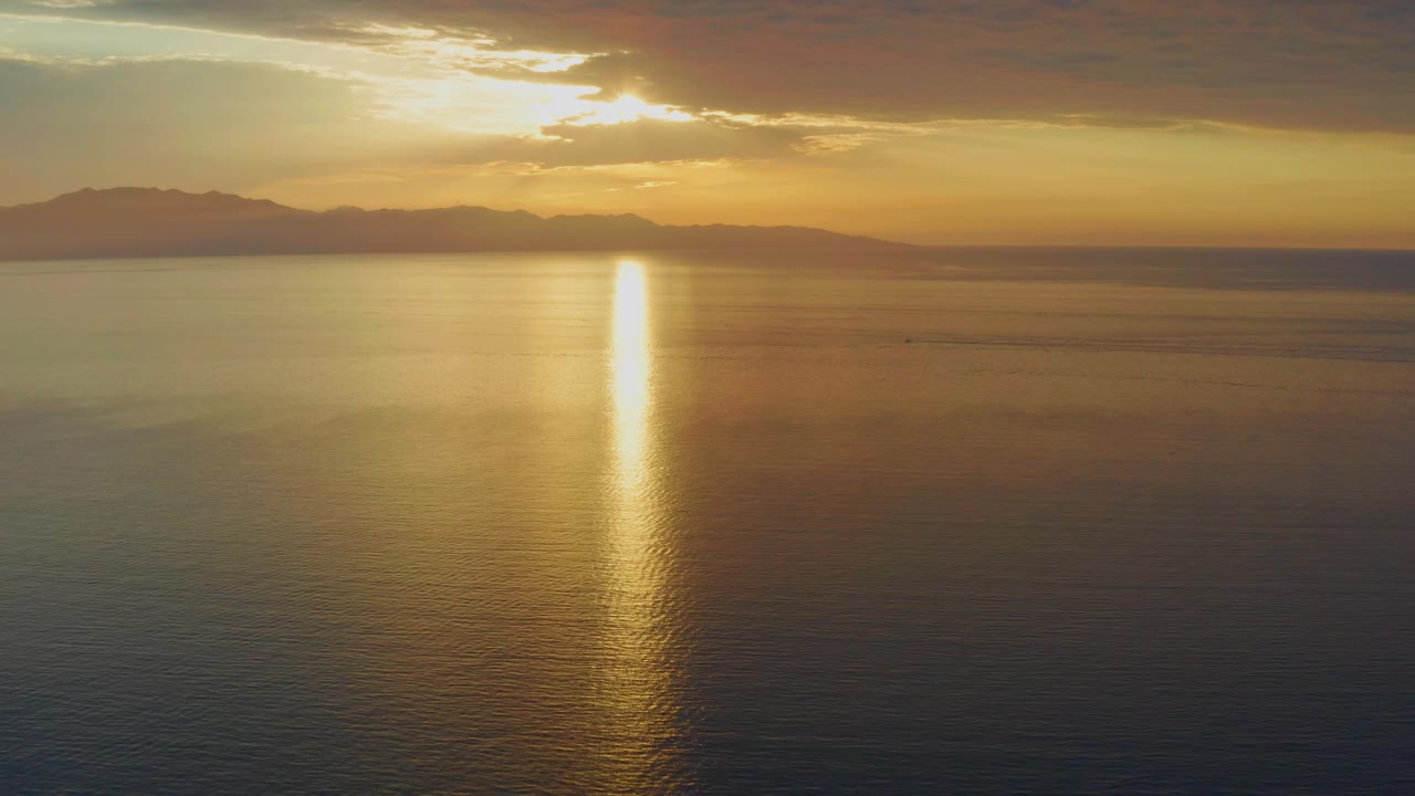 Third frame showing a bright golden reflection on darker ocean water, low mountains across the background, orange cloud cover, and no visible people in the scene.