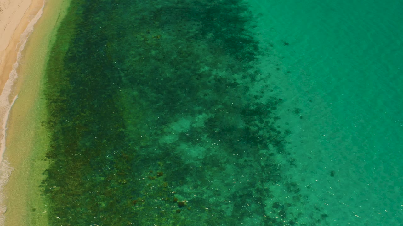 Aerial beach edge beside dark underwater patches and clear turquoise water.