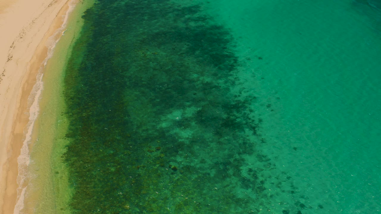 Opening overhead frame of El Sargento La Ventana shoreline, with golden sand at left, turquoise shallows at right, dark underwater patches near the beach, and no visible people.