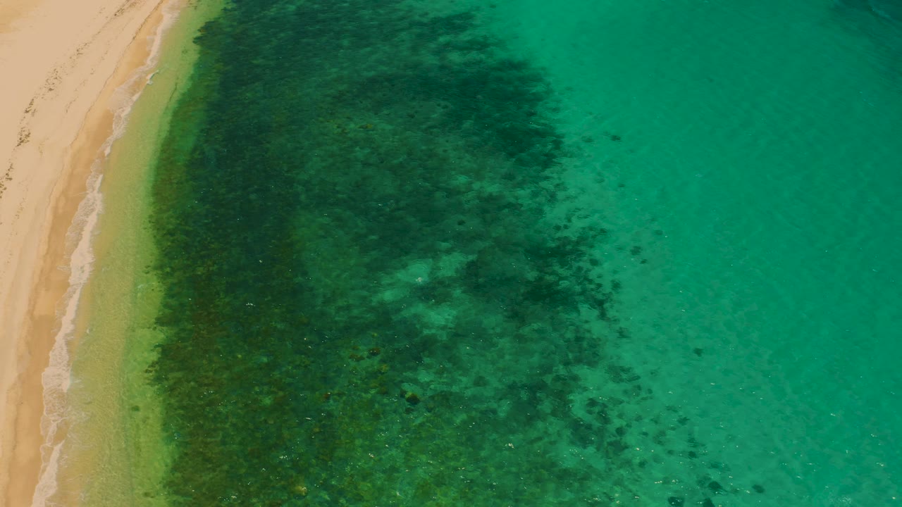 Overhead view in El Sargento La Ventana with the sand strip still on the left, a narrow pale surf line, green shallows, darker seafloor areas, and no visible people.