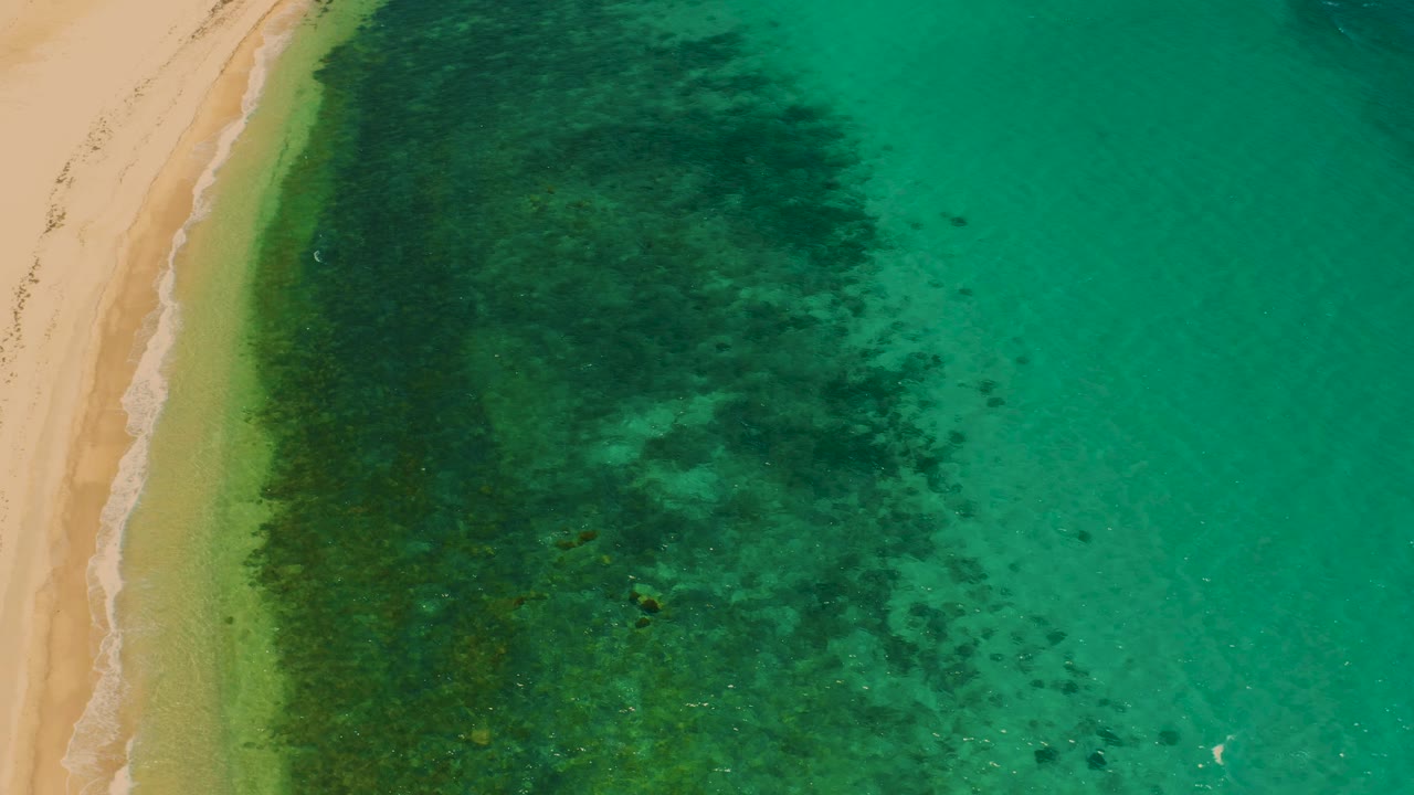 Top-down El Sargento La Ventana beach frame showing more open green water to the right, the sand and foam line at left, dark underwater shapes, and no visible people.