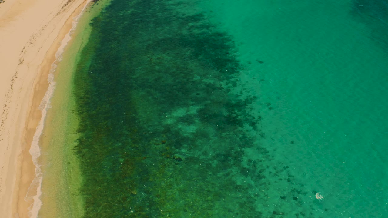 Mid-clip overhead view of El Sargento La Ventana coast, with a broad dark underwater patch centered beside the sand, clear green water, faint surf, and no visible people.