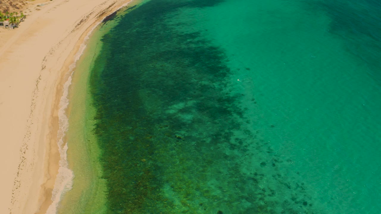 High aerial frame over El Sargento La Ventana, Mexico, showing a curved sandy shore on the left, green shallows, darker underwater patches, and no visible people.