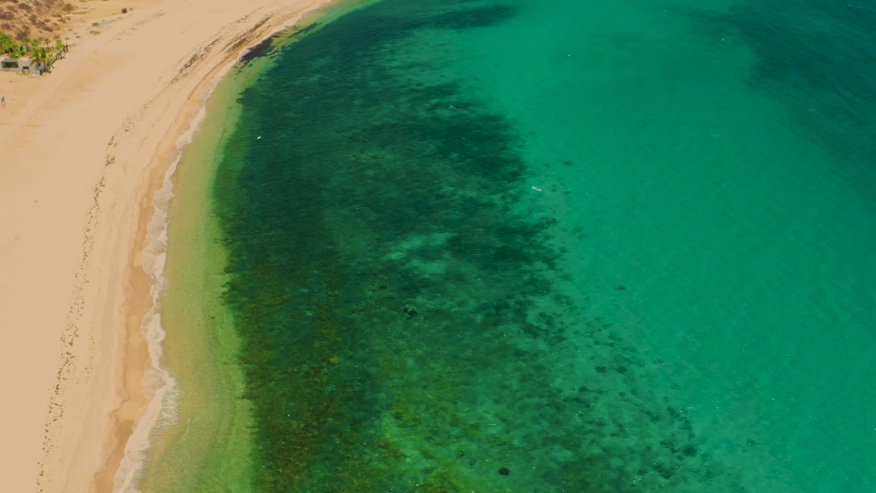 High aerial frame from El Sargento La Ventana, Mexico, showing pale sand at left, a green band of shallow water, darker seabed texture, and open turquoise sea with no visible people.