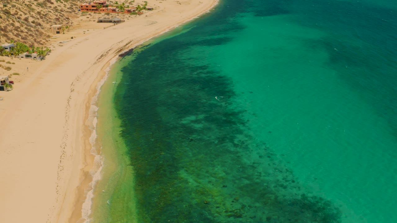 High aerial view in El Sargento La Ventana, Mexico, showing the beach running down the left side, turquoise water on the right, darker reef or seaweed texture, and no visible people.