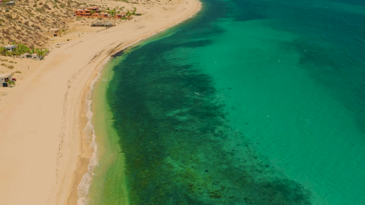 Aerial coastal frame over El Sargento La Ventana, Mexico, with empty-looking sand at left, green water filling most of the frame, a dark underwater patch near center, and no visible people.