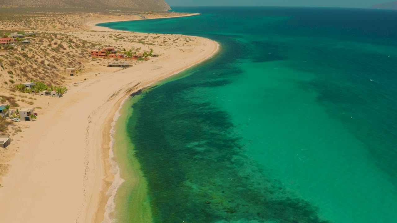 Elevated view over El Sargento La Ventana, Mexico, with green-blue shallows along the beach, darker underwater areas offshore, desert scrub inland, and no visible people.