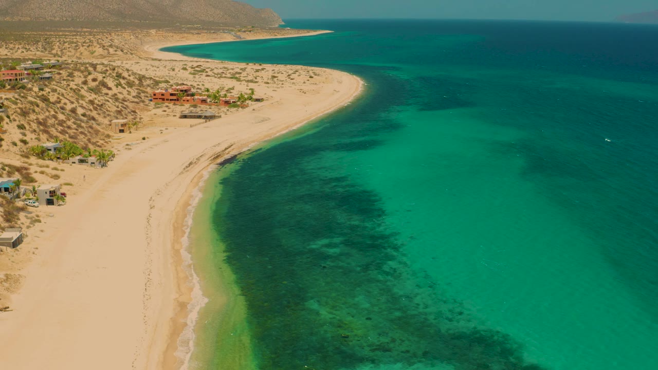 Wide aerial frame of El Sargento La Ventana, Mexico, showing the beach curve extending toward distant hills, turquoise water filling the right side, and no visible people on the sand.