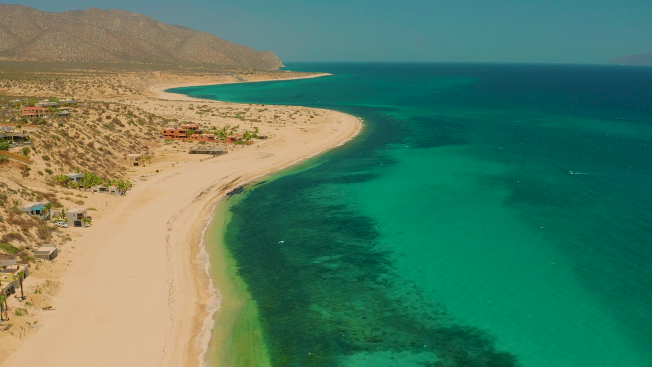 At 0:00, an aerial view over El Sargento La Ventana shows a broad sandy beach curving around clear turquoise water, low desert hills in the background, scattered shore buildings, and no visible people.