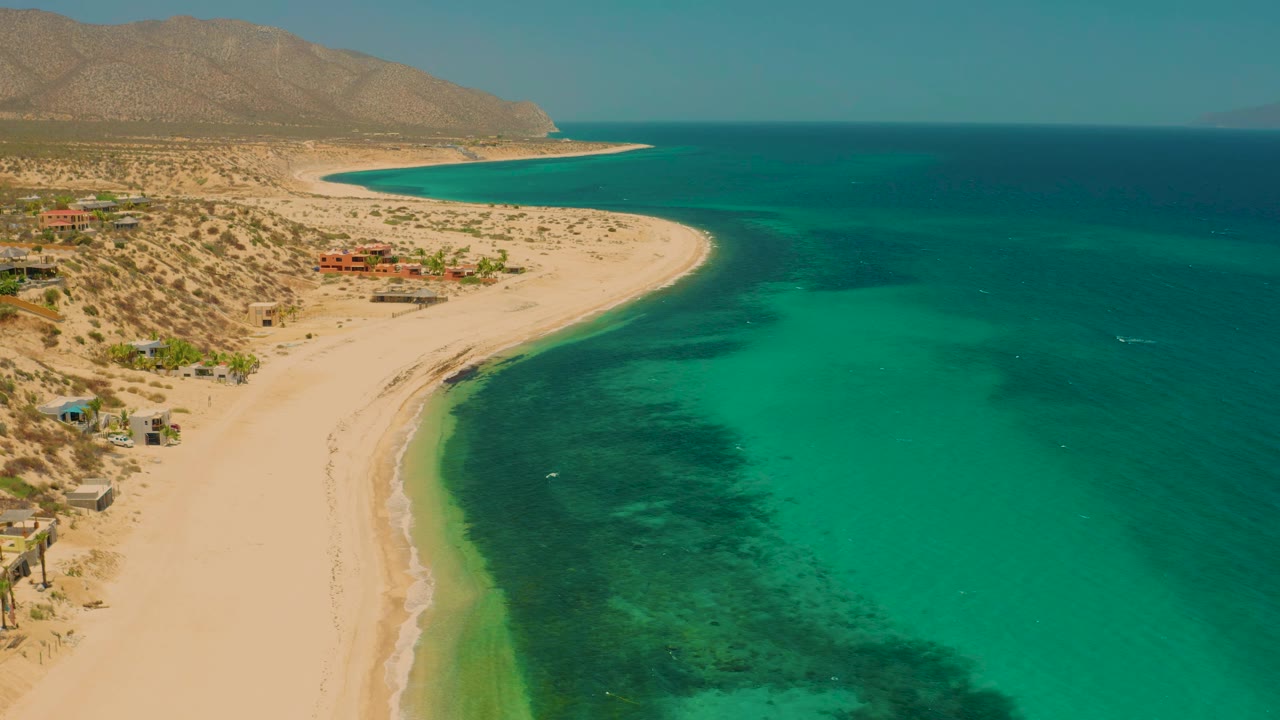 At 0:01, the aerial composition holds on El Sargento La Ventana with the sandbar-like beach edge, clear green-blue shallows, desert hills, small shoreline structures, and no visible people.