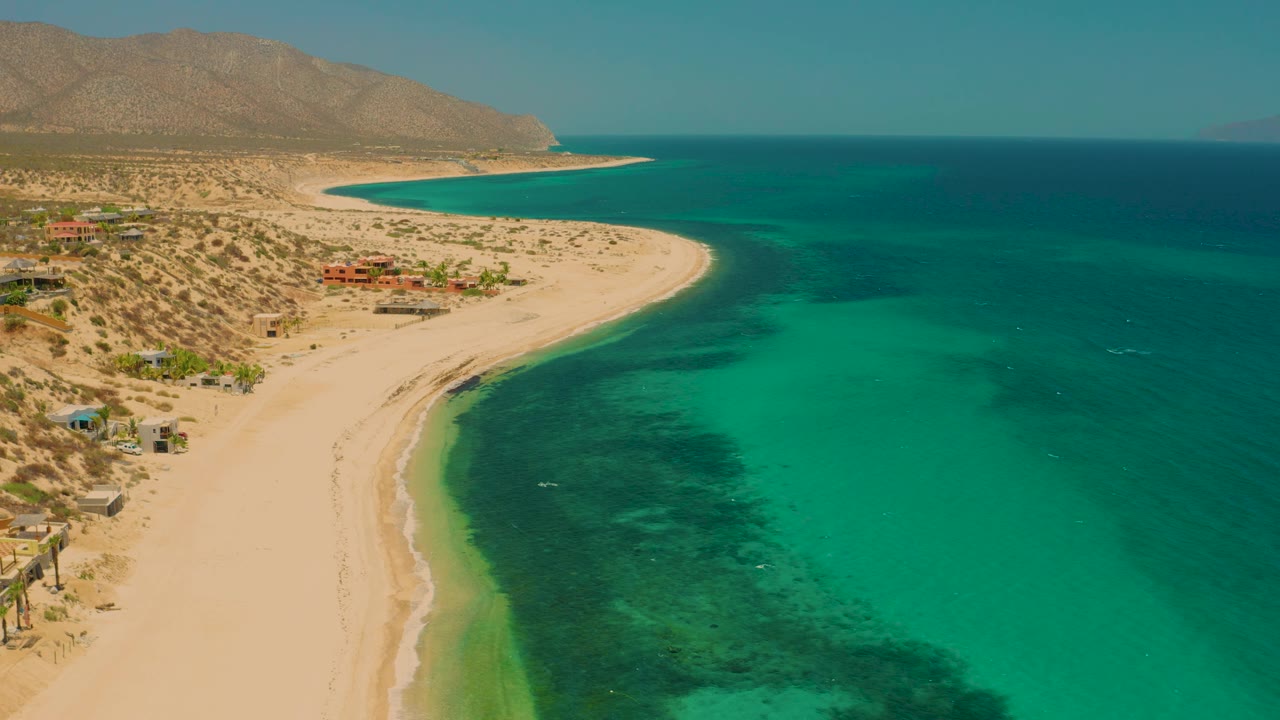 At 0:02, El Sargento La Ventana is seen from above with more open water filling the right side, the pale beach sweeping along the left, desert hills behind it, and no visible people.