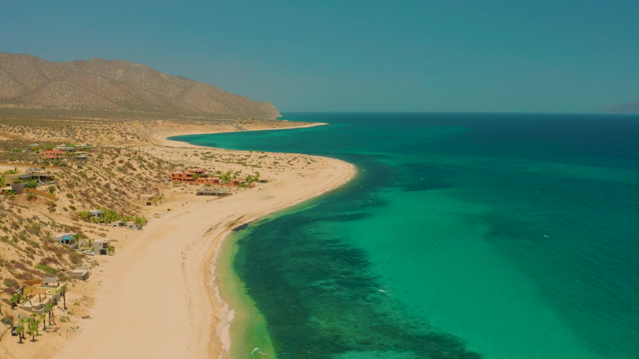 Opening aerial frame over El Sargento La Ventana, Mexico, showing a pale curved beach, turquoise shallows, dry hillside vegetation, scattered small buildings, distant mountains, and no visible people.