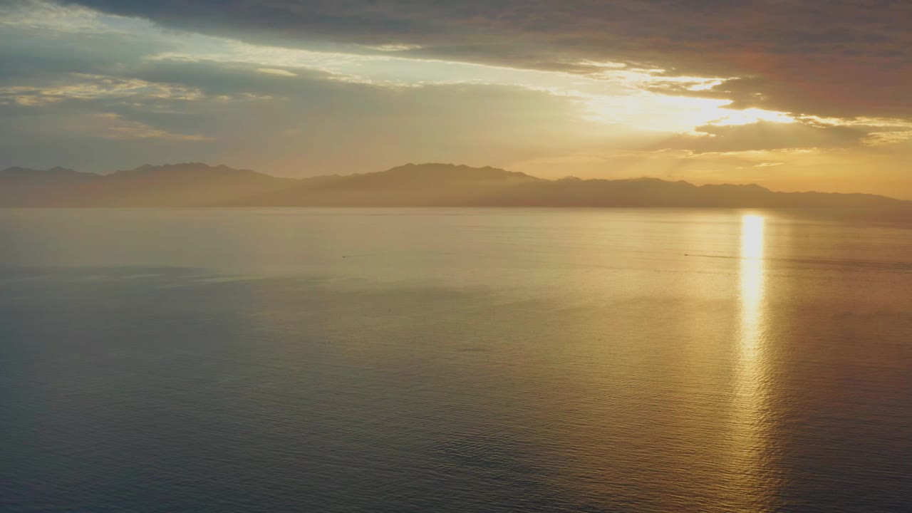 Early frame over El Sargento La Ventana, Mexico, with the sun hidden behind clouds, a narrow gold column reflected on the water, hazy mountains across the horizon, and no visible people.