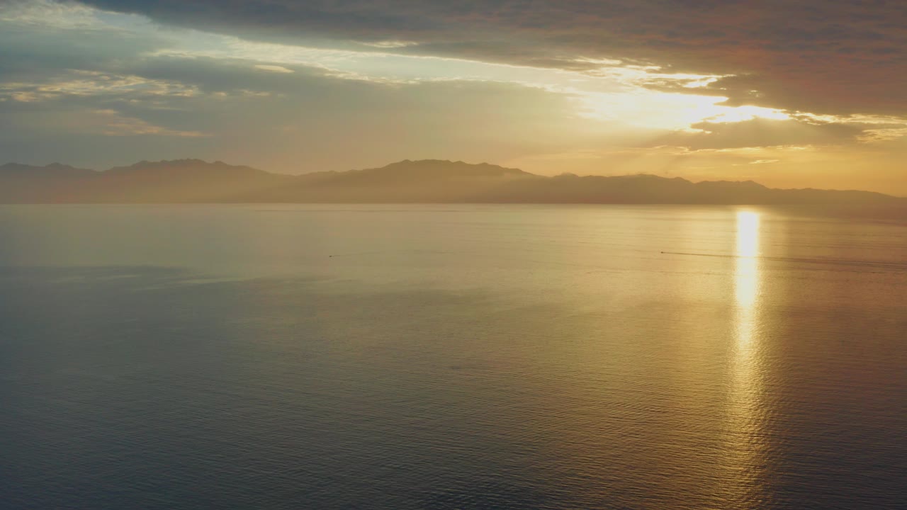 Opening view over water in El Sargento La Ventana, Mexico, with a bright vertical sun reflection, hazy hills, layered clouds, and no visible people.